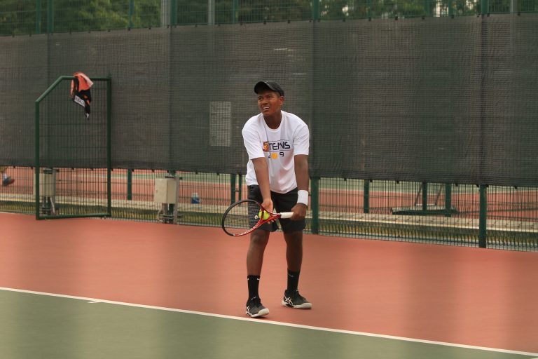 An ACS(I) tennis player prepares to serve during a competitive match, viewed from the opposite baseline.